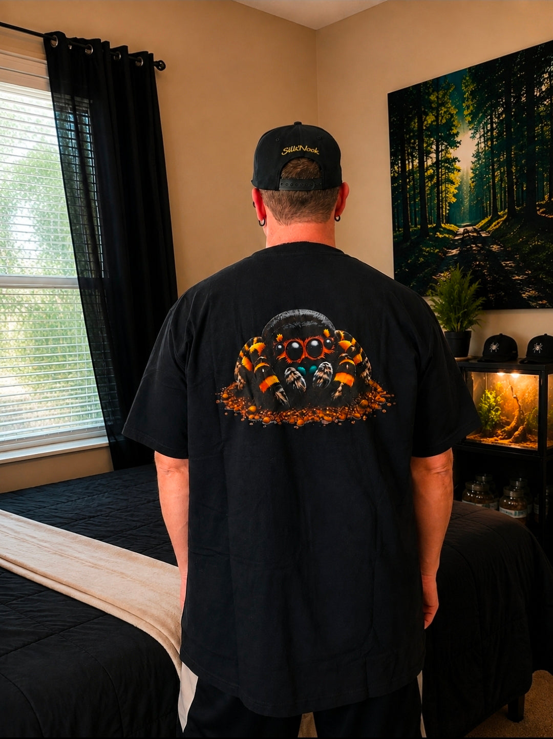 Black SilkNook T-shirt featuring a detailed orange and black jumping spider design on the back, displayed against a warm natural-inspired background.